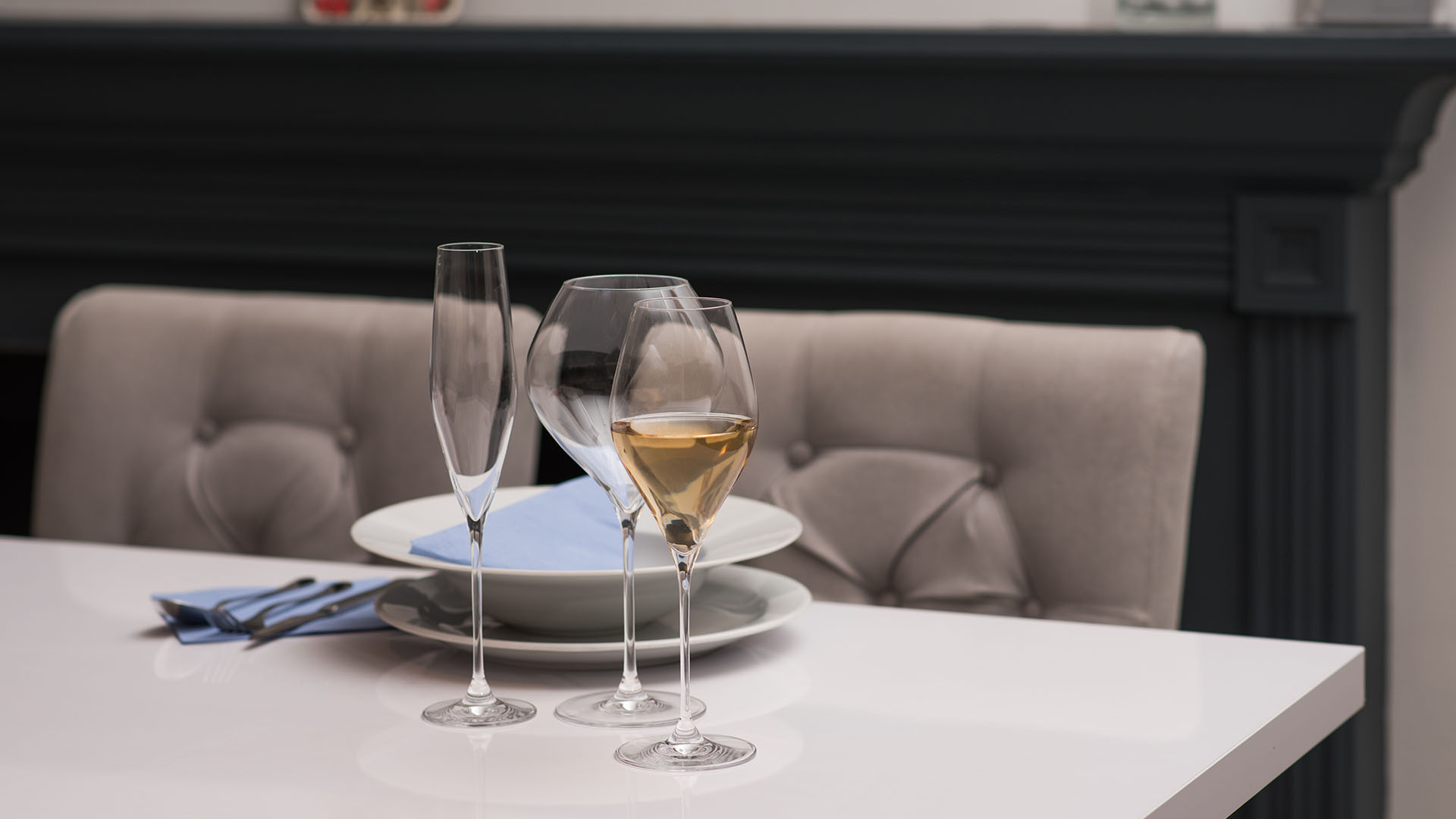 Three Rona stemmed glasses including a champagne flute and two wine glasses arranged on a white table top with a dark fireplace surround in the background